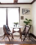 Cozy dining area with a round table, wooden chairs, plants, and framed art on cream walls.