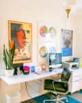 Home office with a desk, computer, printer, black chair, potted plant, and colorful artwork on the walls.