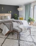 Modern bedroom with a wooden bed, dark accent wall, patterned rug, and plants by a large window with teal curtains.