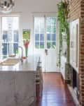 Marble kitchen island with sink, flowers, and stools, next to French doors and a brick wall with hanging plants.