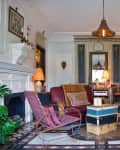 Eclectic living room with vintage decor, leather armchair, wicker chairs, ornate fireplace, and stacked suitcases as a table.