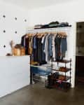Open closet with hanging clothes, white dresser, shoe rack, and a small cart in a room with cross-patterned wall.