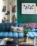 Blue tufted sofa with colorful cushions, floral curtains, and a vase of purple flowers on a coffee table in a green-walled room.