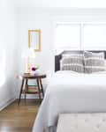 Bright bedroom with a white bed, patterned pillows, side table with lamp, and a hat on the wall.