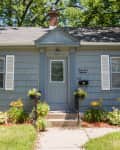 Blue house with white shutters, front door, and flower garden, surrounded by trees.