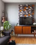 Mid-century modern living room with a TV on a wooden console, patterned wall art, guitar, and a cozy chair with a pouf.