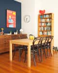 Dining room with wooden table, black chairs, bookshelf, wall clock, and colorful poster on a blue accent wall.