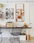 Home office with wire chairs, a long desk, corkboard with notes, abstract art, and a small plant on a shelf.
