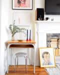 Small desk with eucalyptus vase, blue candles, and framed art near a fireplace with stacked books and a TV above.