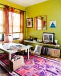 Home office with lime green walls, pink patterned rug, desk with red lamp, and art on shelves and walls.