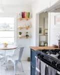 Bright kitchen with wooden countertops, black cabinets, round table, white metal chairs, and wall shelves with books and plants.