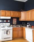 Kitchen with wooden cabinets, navy walls, white stove, and dishwasher. Decor includes framed art and colorful mugs.