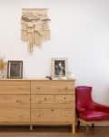 Wooden dresser with framed photos, macramé wall hanging, red chair, and potted plants by a window.