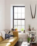 Bright living room with yellow sofa, NYC pillow, wooden desk, plants, and large windows.