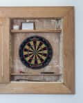 Wooden dartboard cabinet with open door, displaying a dartboard, darts, and a small shelf with a baseball and framed item.