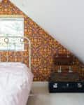 Attic bedroom with floral wallpaper, white metal bed, and stacked vintage suitcases.
