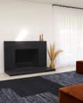 Modern living room with black brick fireplace, brown sofa, beige chair, pampas grass, and a dark patterned rug.