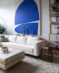 Bright living room featuring a white sectional Sabai sofa, large plants, a blue abstract painting, and a round coffee table.