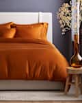 bedroom featuring Quince products: an orange duvet set, white upholstered headboard, and a side table with a lamp and decorative vase.