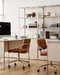 Modern office space featuring two desks with computers, stylish brown chairs, and a metal shelving unit with decor items.