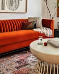 Living room featuring an orange velvet sofa, round coffee table, books, a glass of water, and potted plants.