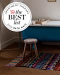 A blue freestanding bathtub with a wooden tray, a small wooden stool, and a colorful patterned rug on a woven floor.
