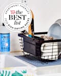 Black dish drying rack full of dishes and utensils sitting on a kitchen counter next to the sink. There is a seal in the upper left corner that reads "Apartment Therapy the Best List 2023 Dish Drying Racks"