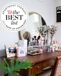 Wood vanity table with makeup in organizers, round mirror, plants around. Seal in upper left reads "Apartment Therapy the Best List 2024 Makeup Organizers"