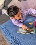 A child in a purple shirt plays on a blue blanket, interacting with a red toy speaker and a storybook.