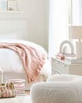 Cozy bedroom featuring a white bed with a pink knitted throw, a small stool, a woven bag, and a plush chair.