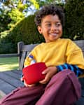 A smiling child in a yellow shirt and striped sleeves sits on a wooden bench, holding a red speaker with a toy figure.