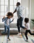 Family dancing in a living room with a gray sofa, colorful rug, and large windows.