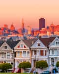 Row of colorful Victorian houses with San Francisco skyline at sunset.