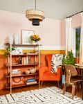 Colorful home office with orange chair, wooden desk, laptop, record player, and bookshelves. Cream curtains and plants add decor.