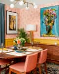 Dining room with pink chairs, round table set for four, floral wallpaper, blue curtains, and vibrant artwork on walls.