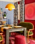 Eclectic dining room with green velvet chairs, geometric wallpaper, wooden table, and orange pendant light.