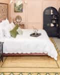 Side view of a pink velvet bed frame with white scalloped duvet. The room has pink walls, a black armoire and side table, and a gold framed standing mirror.