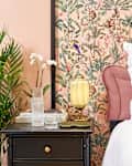 Corner view of the left side of the bed with a pink headboard, a green velvet throw pillow, and a black bedside table with a carafe and cup of water, and a white orchid. Anthropologie Eloise table lamp.
