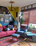 Eclectic living room with red sofa, blue armchair, colorful wall art, plants, and a small round table by the window.