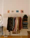 Clothing rack with colorful garments, stacked clothes, desk with mirror, and framed art on walls in a cozy room.
