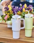 Two insulated mugs with straws on a wooden table, next to a colorful flower arrangement outdoors.