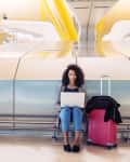 Woman sitting on airport floor with laptop, next to pink suitcase and jacket, against a modern metallic wall.