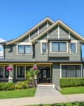 Two-story gray house with white trim, front porch, manicured lawn, and hanging pink flowers under a clear blue sky.