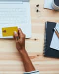 Hands typing on a laptop keyboard while holding a yellow credit card, with notebooks and a cup of coffee nearby.
