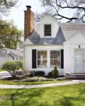 White cottage-style house with black shutters, brick chimney, and manicured lawn, surrounded by trees.