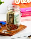 Mason jar with cash and coins on a wooden tray, next to sunglasses and a vase, with colorful books in the background.