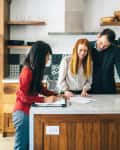 Three people reviewing documents at a kitchen island with wooden cabinets and modern appliances.