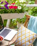 Laptop on a wooden table with flowers, a bowl of strawberries, and a patterned pillow on a balcony.