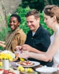Group of friends enjoying an outdoor meal at a wooden table with flowers, wine, and salads.