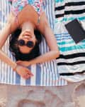 Woman in sunglasses lying on a striped beach towel, smiling on sandy beach.